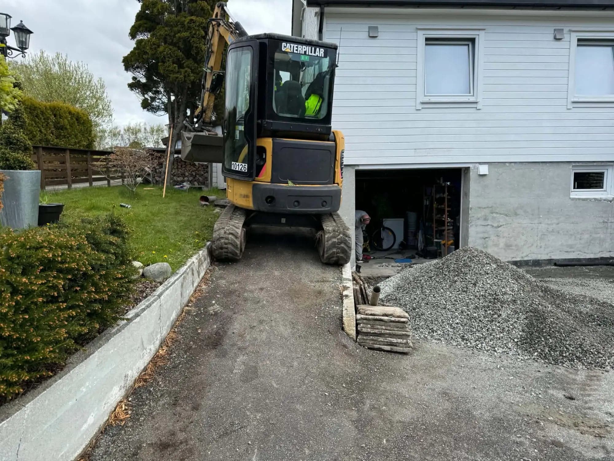 Caterpillar minigraver i smal asfaltert innkjørsel mellom kantstein og hvitt hus, med pukkhaug og drensrør foran garasjen