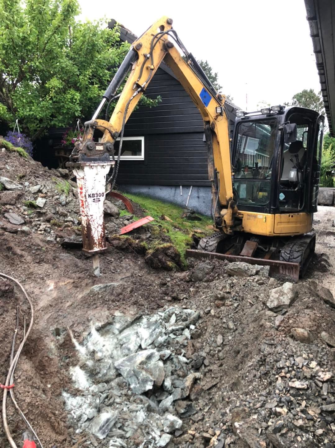 Caterpillar gravemaskin med hydraulisk hammer bryter fjell inntil grunnmuren på et bolighus