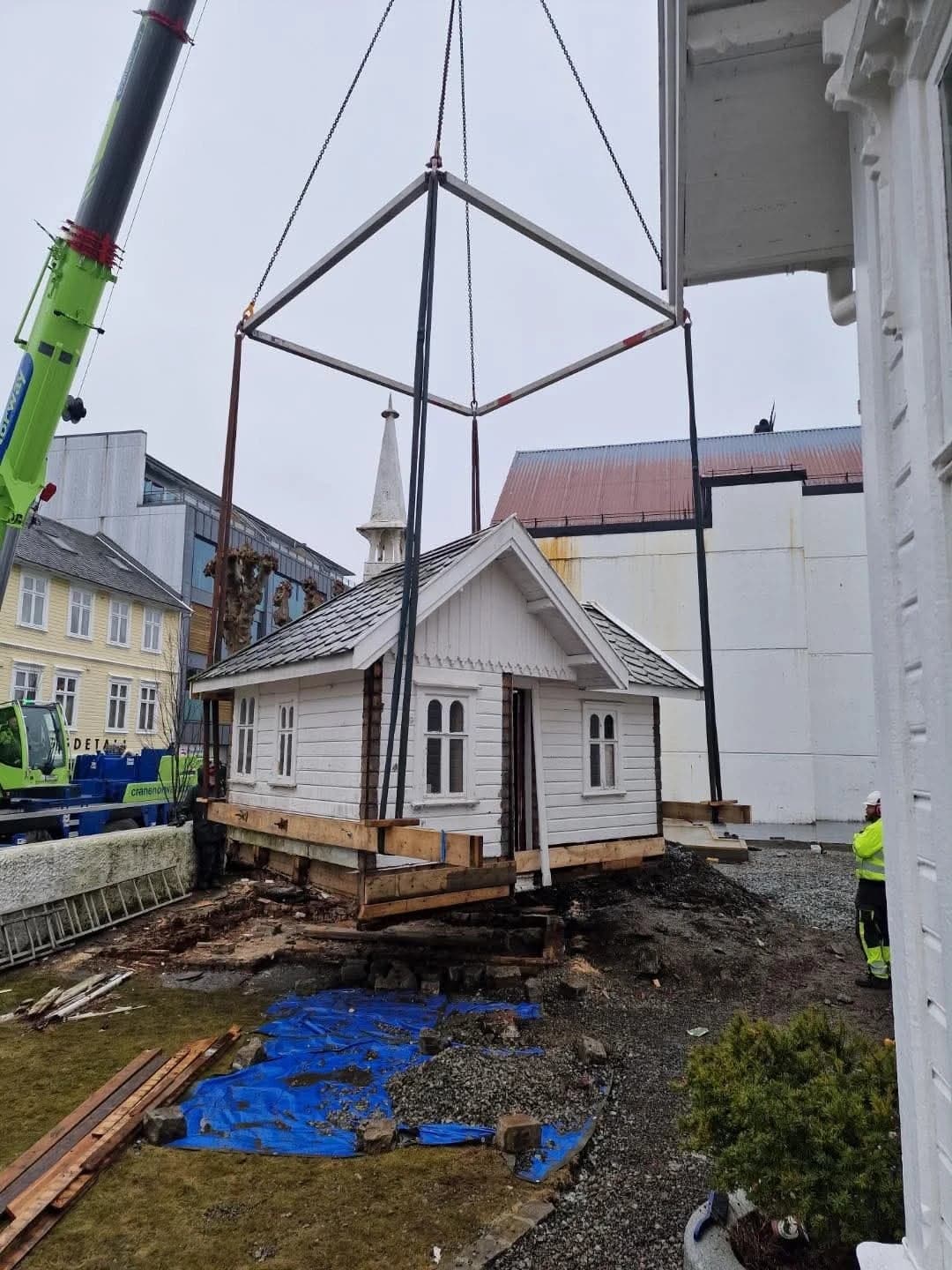 Lite hvitt trehus med spir henger i løfteramme under mobilkran over en grusplass mellom bygårder