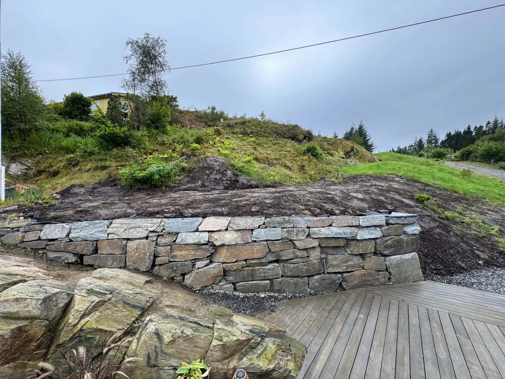 Ferdig tørrmur i naturstein foran en grønn skråning, med pukk ved foten og terrassebord i forgrunnen